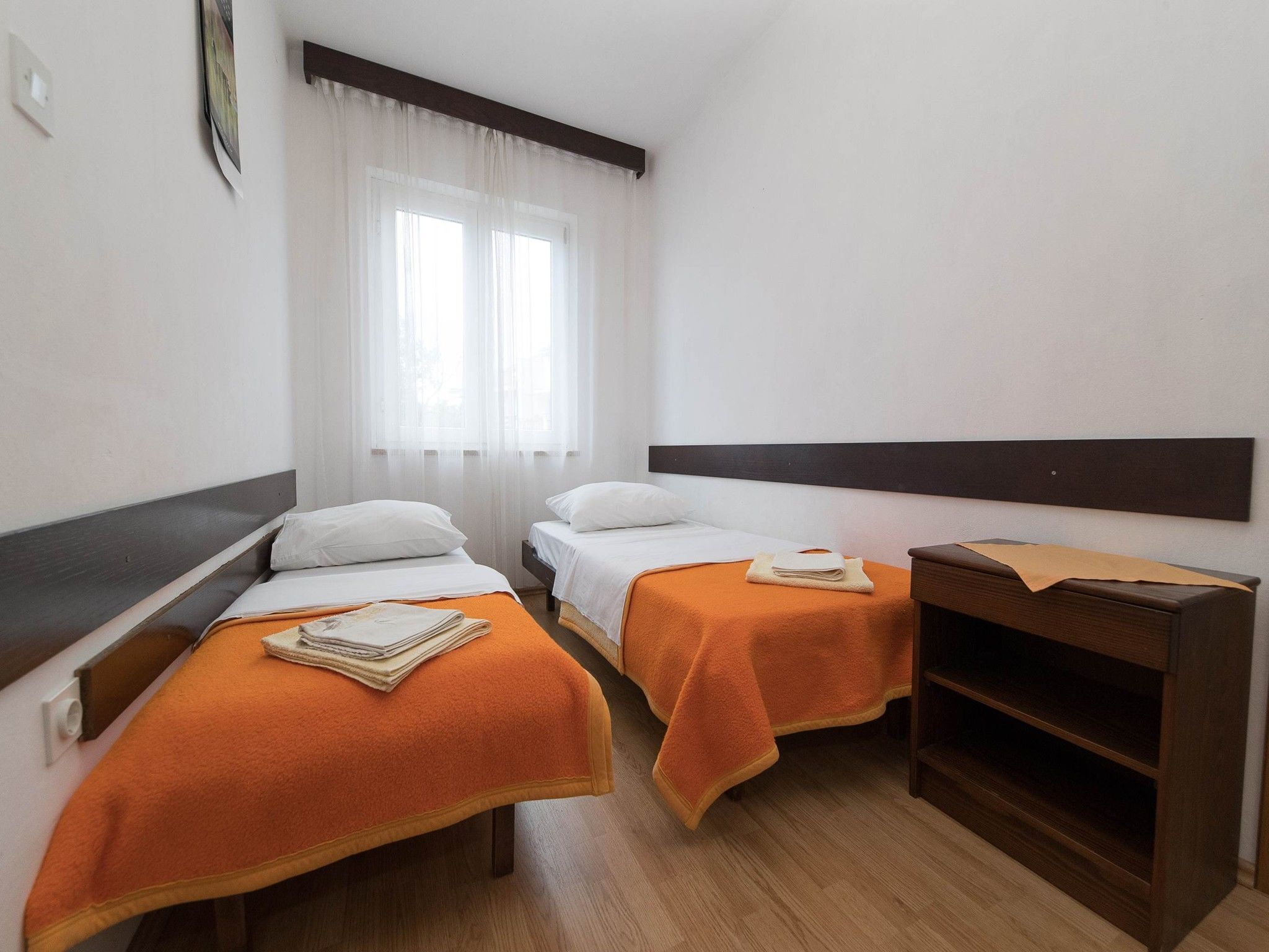 Camera d'albergo con due letti singoli, un comodino in legno, una piccola mensola in legno e una finestra con tende leggere.