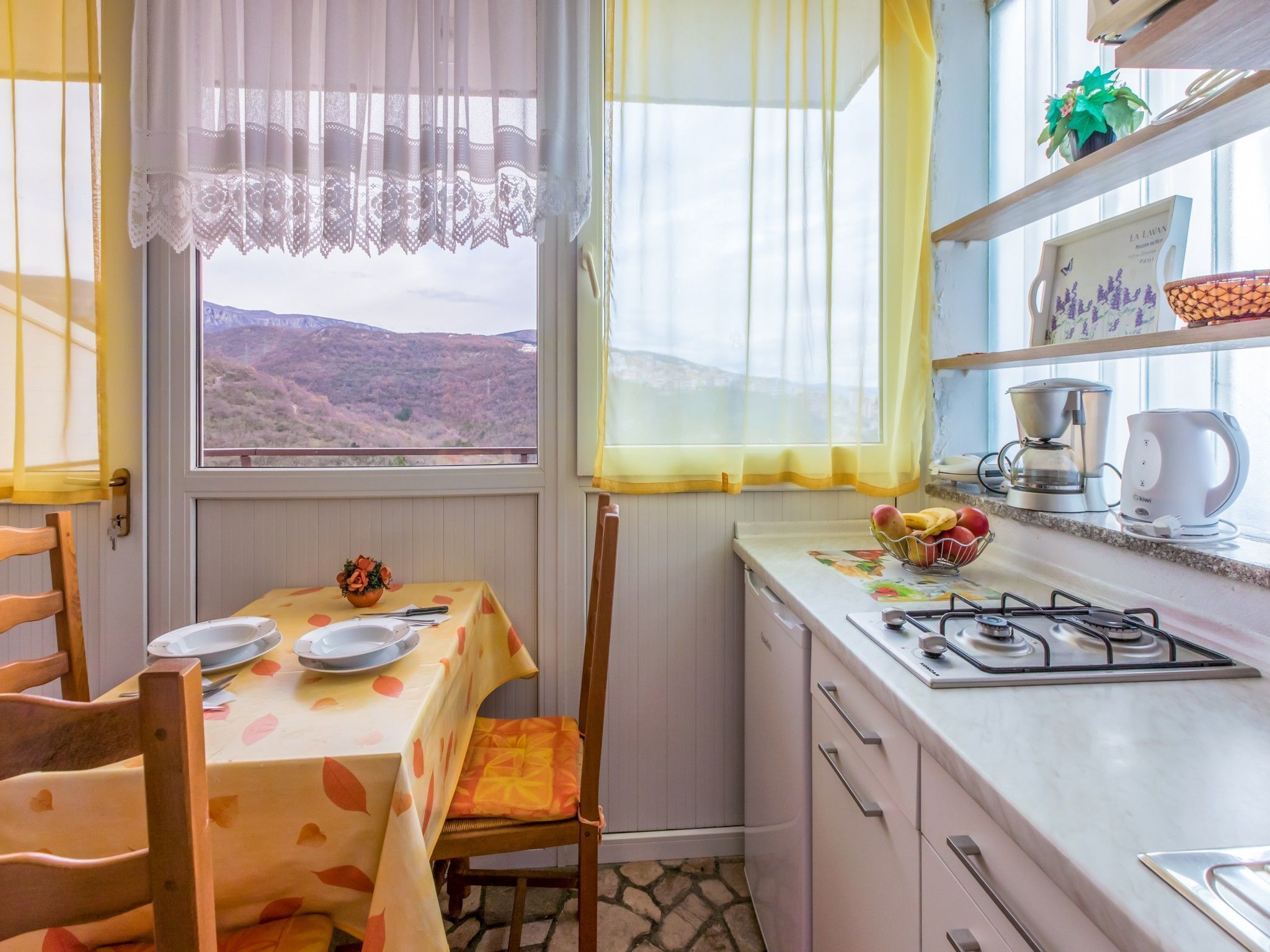 A kitchen with a table set for two, a gas stove, and a view of a mountainous landscape through the window.