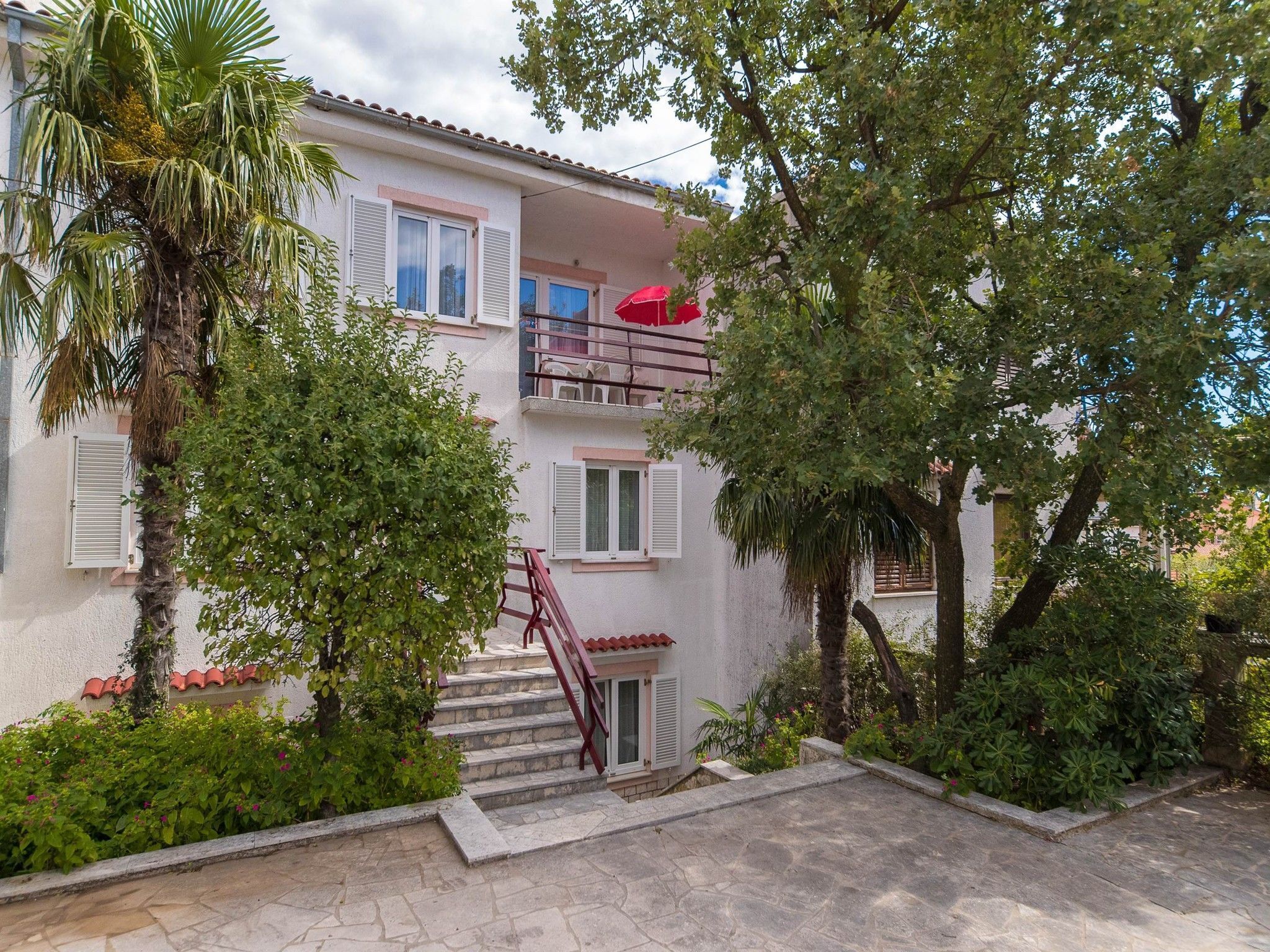 Un edificio a due piani con un balcone, scale che conducono all'ingresso e vegetazione circostante.