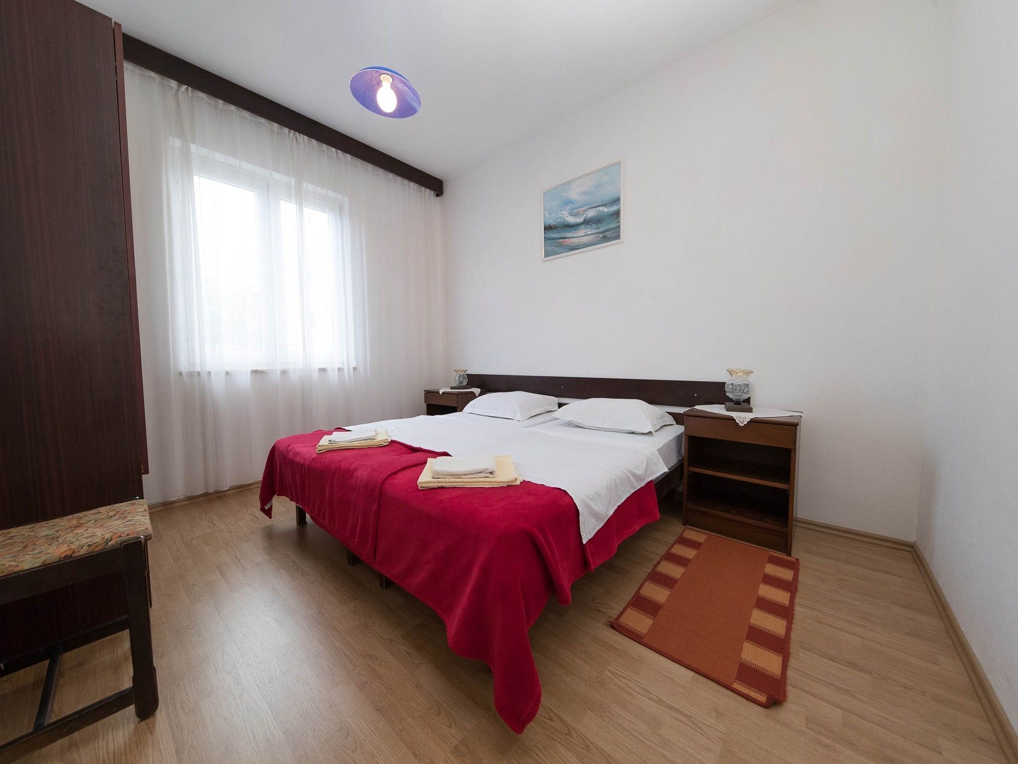 A bedroom with a bed, a bedside table, a nightstand, a window with curtains, a painting on the wall, and a wooden floor.