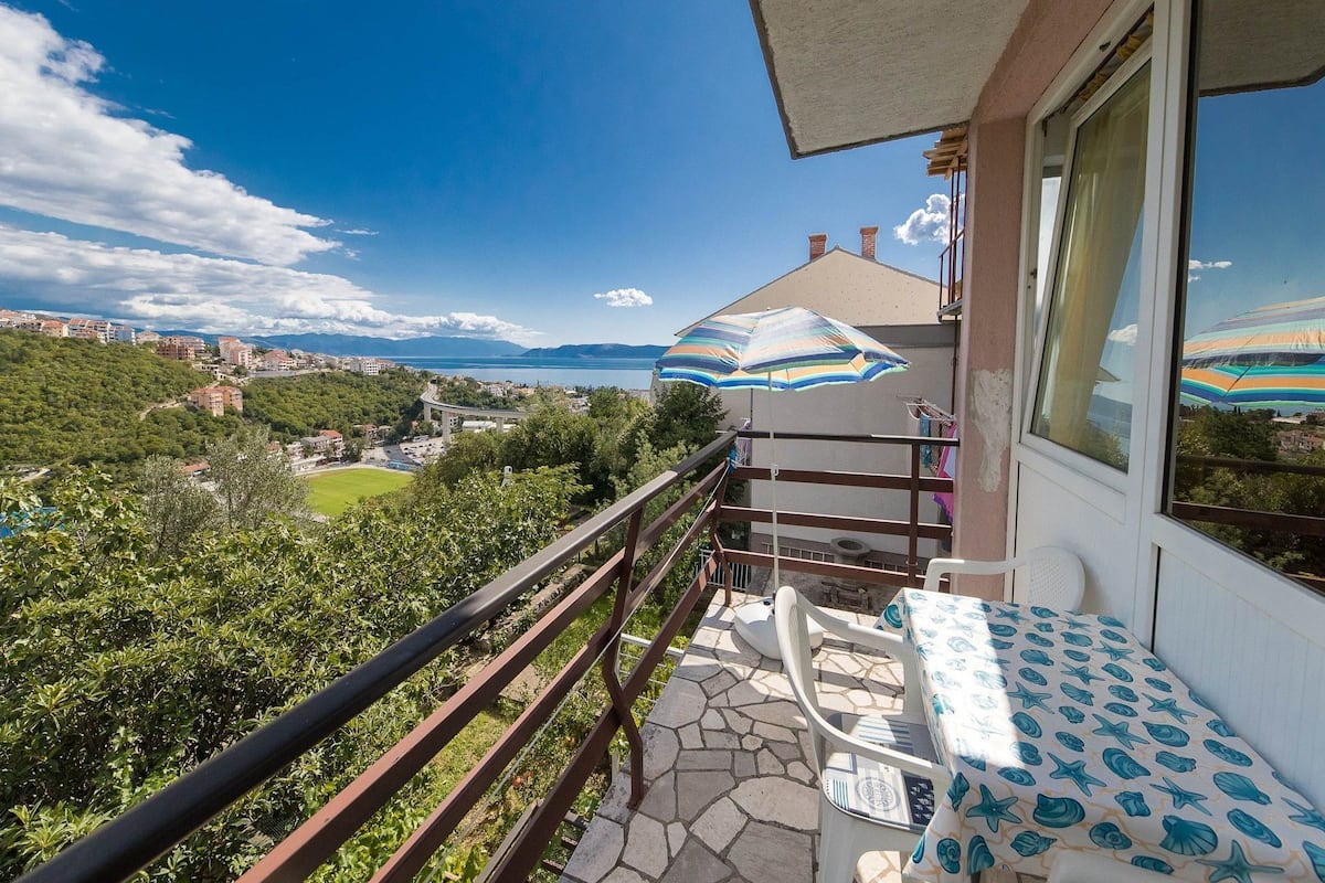 A balcony with a table and chairs, a view of a cityscape and mountains, and a large umbrella.
