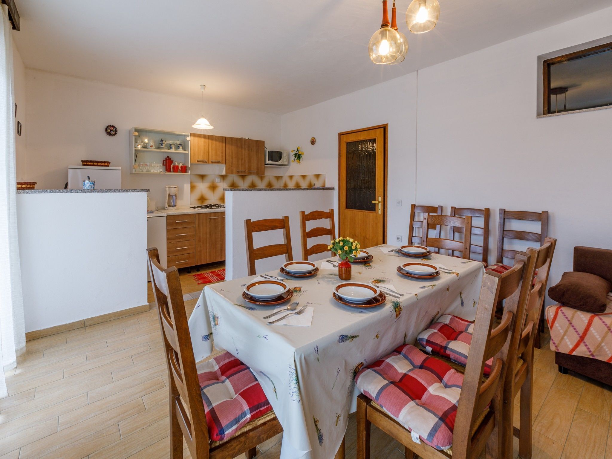 Uma sala de jantar com uma mesa posta para seis pessoas, cadeiras de madeira com almofadas xadrez, cozinha ao fundo e iluminação pendente.