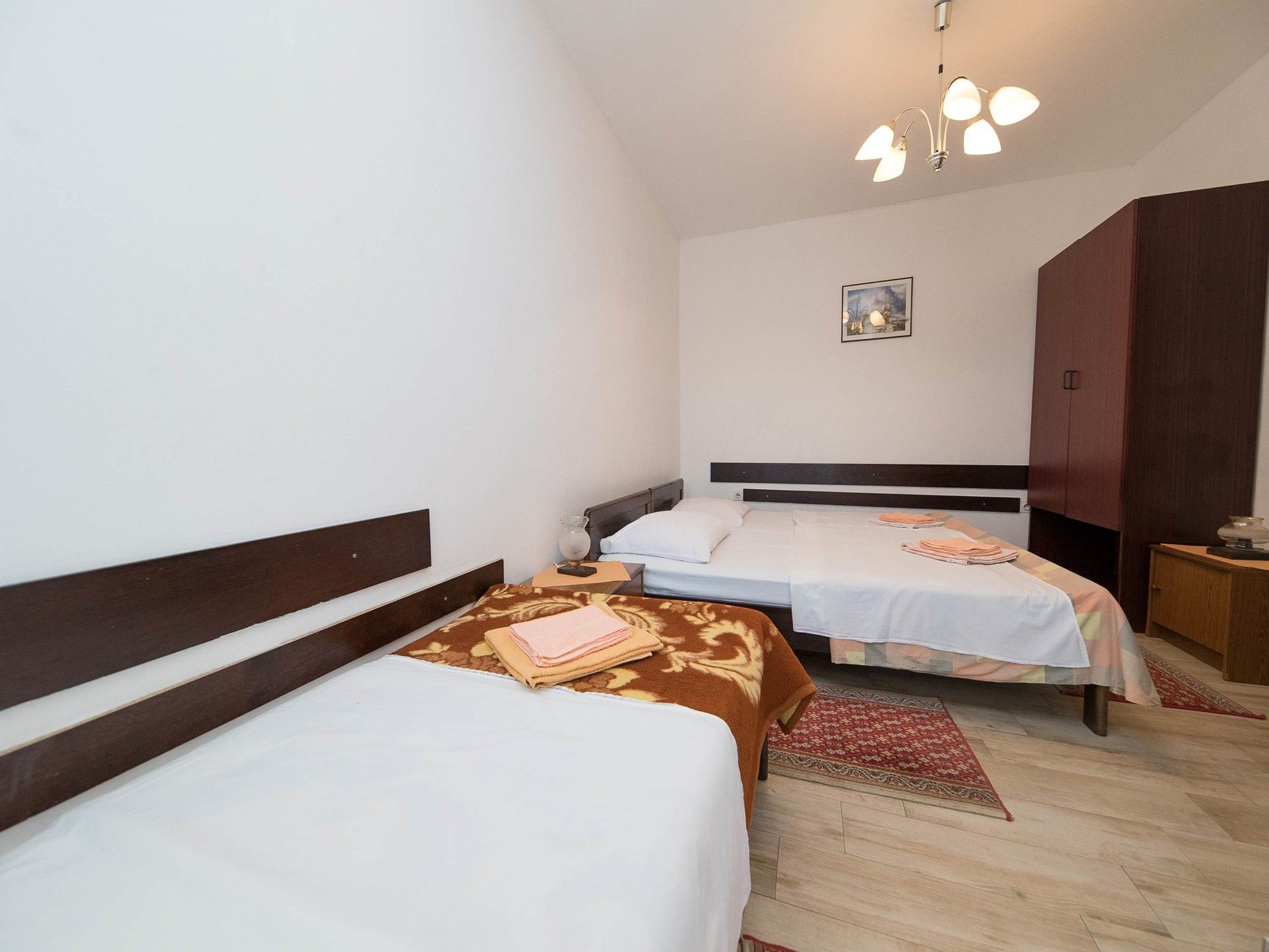 A hotel room with two beds, a wooden wardrobe, a small table, and a ceiling light fixture.
