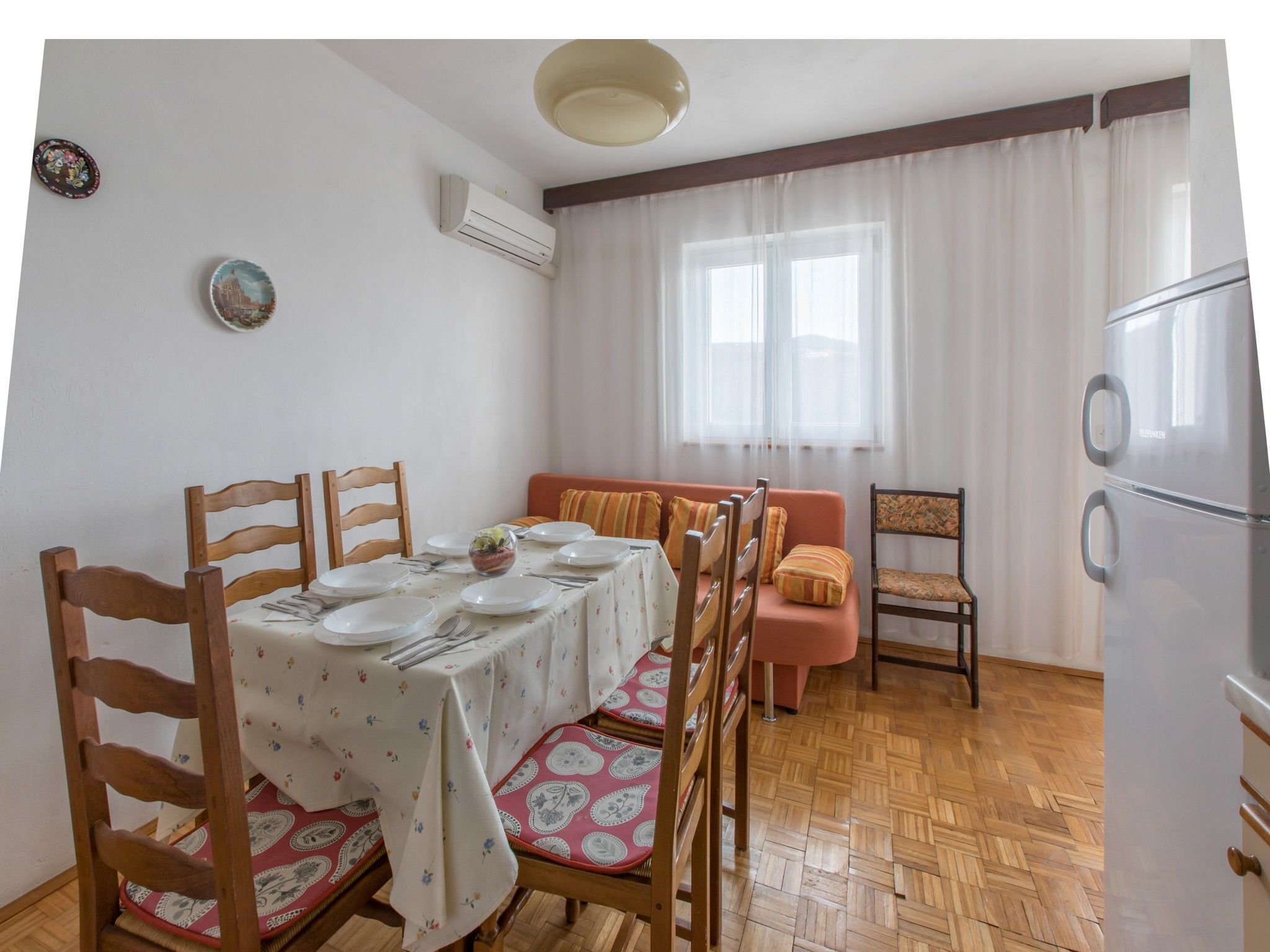 A dining area with a table set for four, wooden chairs, a sofa, a refrigerator, and a window with curtains.