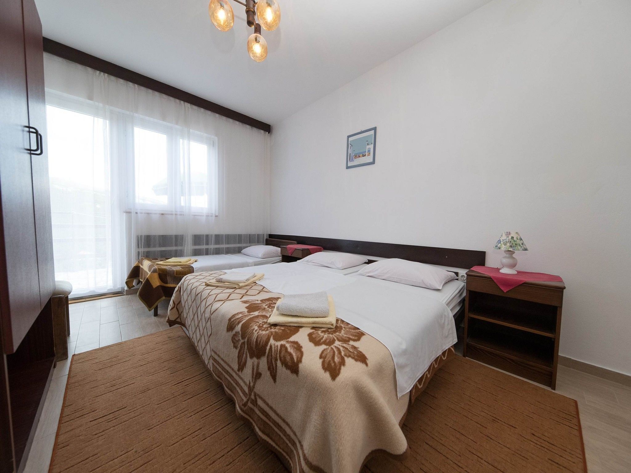 A hotel room with a large bed, two bedside tables, a nightstand, a window with sheer curtains, and a ceiling light fixture.