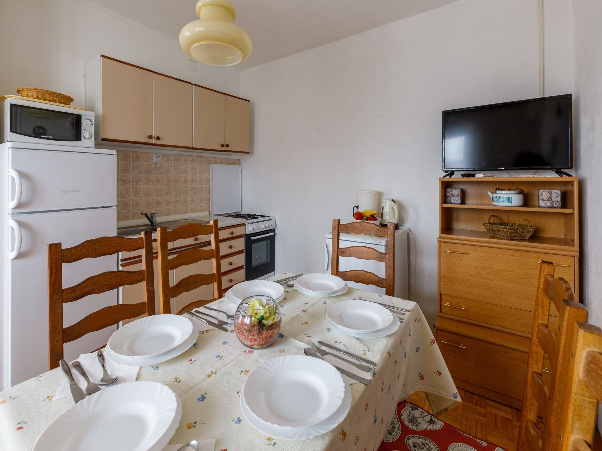 A compact kitchen with a microwave, stove, and refrigerator. There is a dining table set with plates, cutlery, and a decorative centerpiece. A television is mounted on the wall above the table.