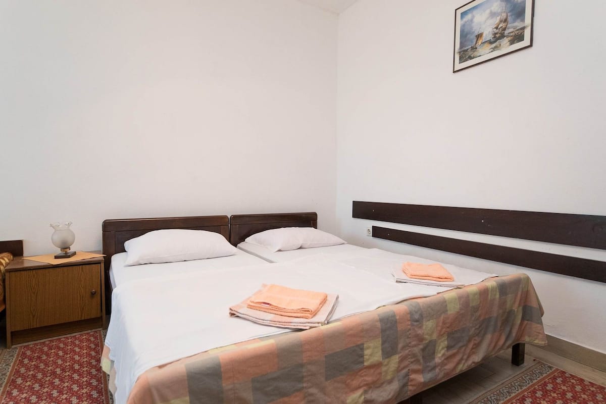 A single bed with a wooden headboard, white bedding, and a checkered blanket. There is a nightstand with a lamp and a framed picture on the wall above the bed.