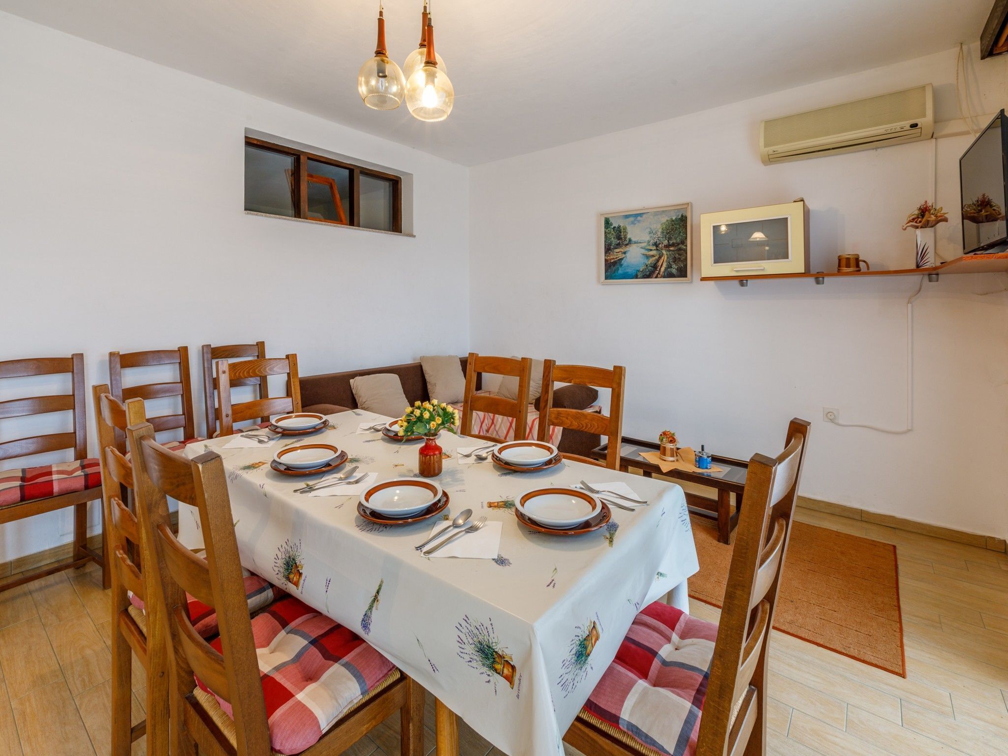 Zona pranzo con tavolo apparecchiato per quattro, circondato da sedie di legno con cuscini a quadri. Sul tavolo, un piccolo vaso con fiori e mensole a parete con oggetti decorativi.