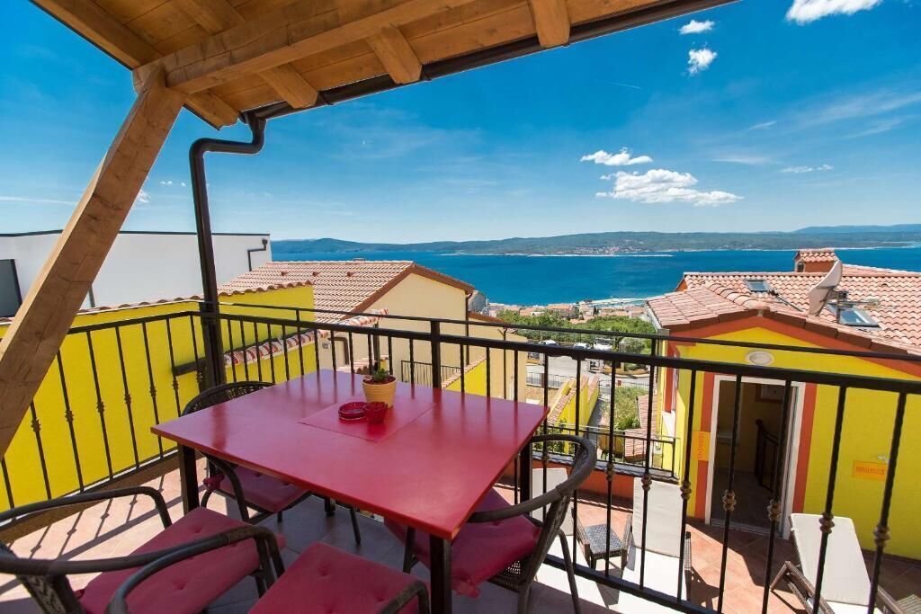 Un balcon avec une table et des chaises rouges, offrant une vue sur la côte, avec des bâtiments et la mer.