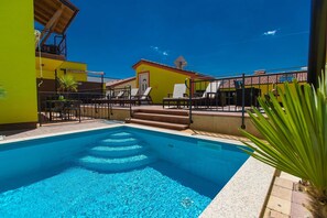 A swimming pool with steps leading into it, surrounded by lounge chairs and a building with a red roof.