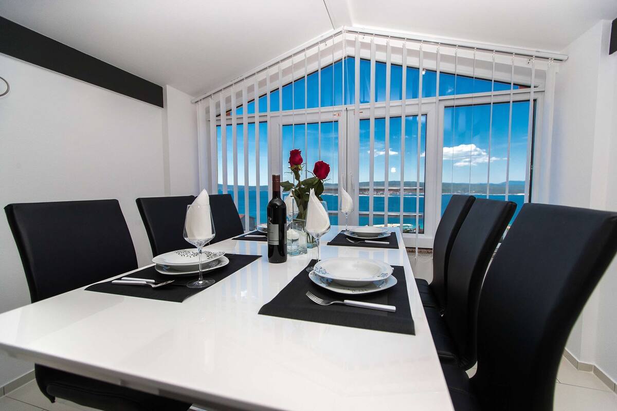 A dining area with a table set for four, black chairs, a vase with red roses, and a view of the sea through large windows.