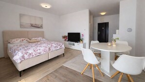 Quarto moderno com cama, televisão, mesa de jantar com cadeiras e pequena cozinha.