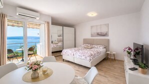 Quarto com cama, uma mesinha com um vaso, uma cadeira, televisão e varanda com vista para o mar.