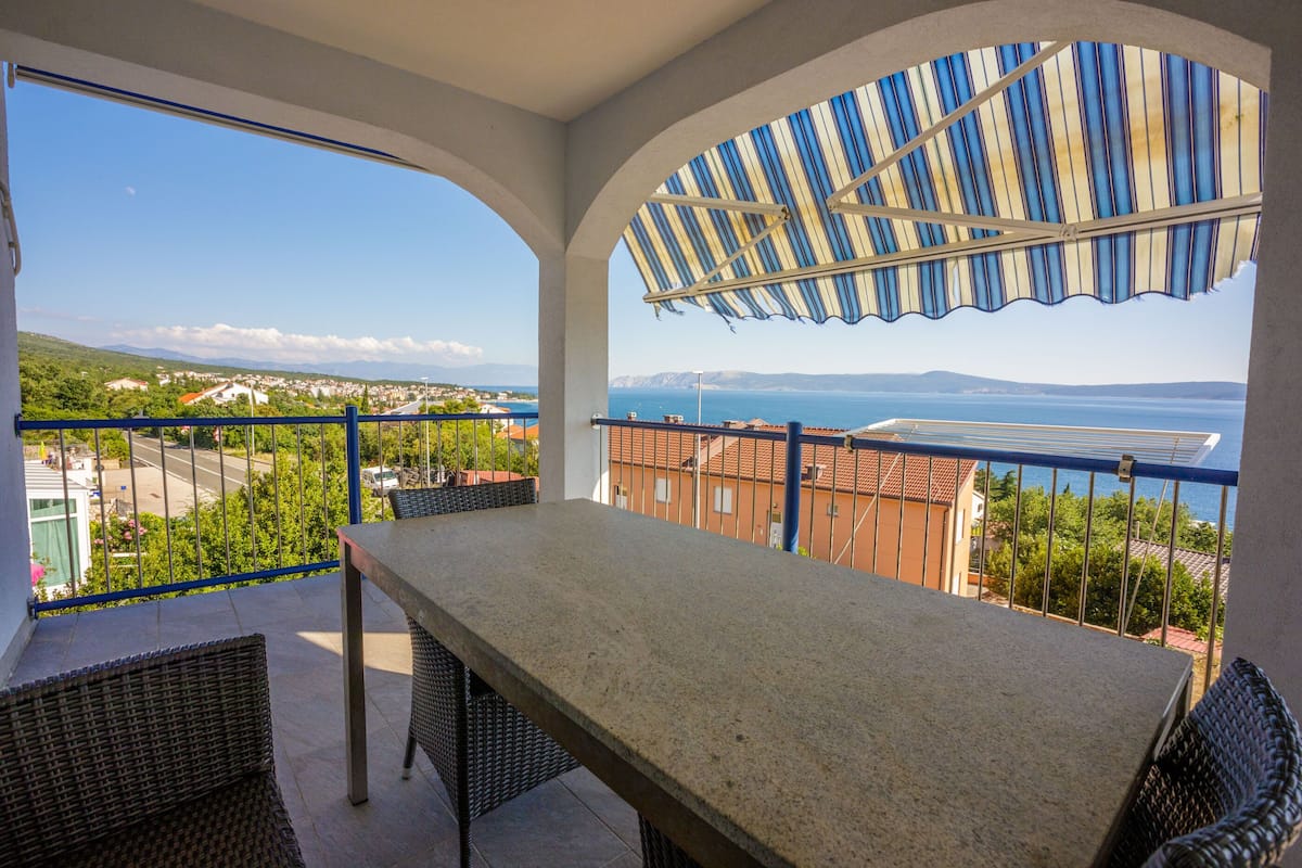 A balcony with a table and chairs, offering a view of the sea and surrounding buildings.