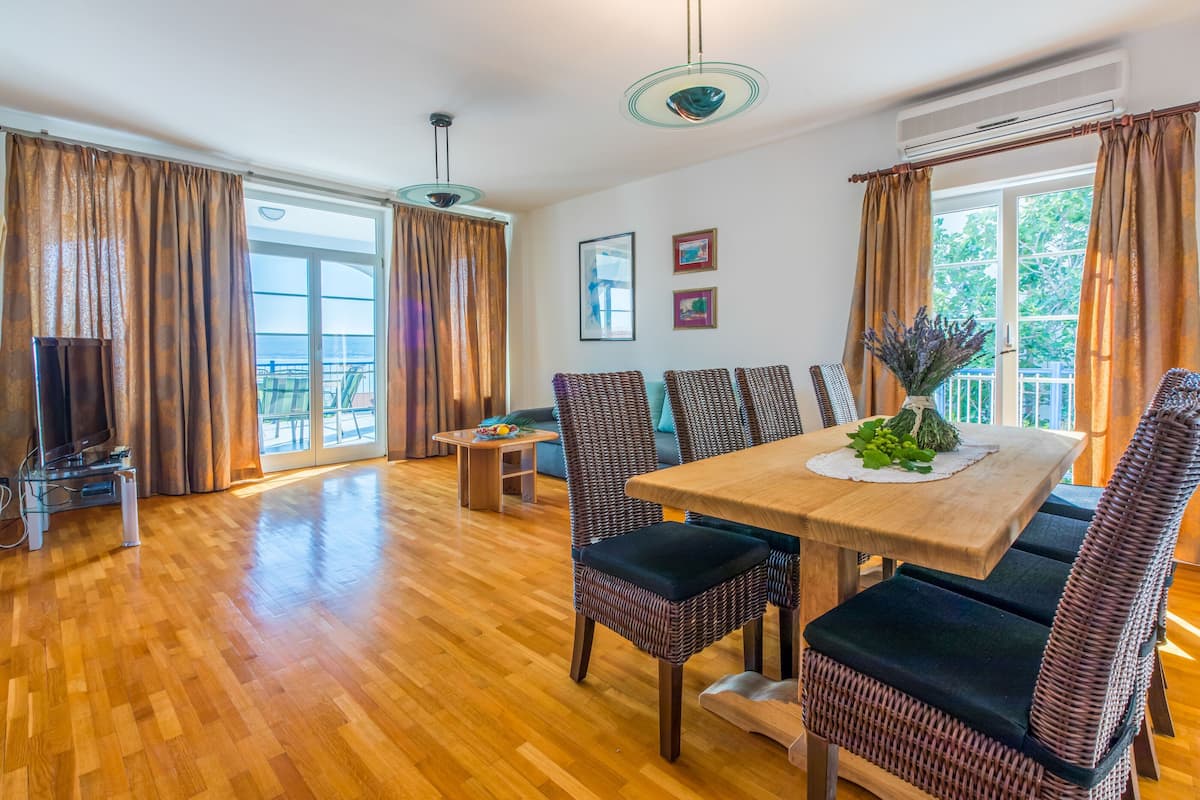 A dining area with a wooden table and wicker chairs, a television, and a view of the outdoors through large windows.