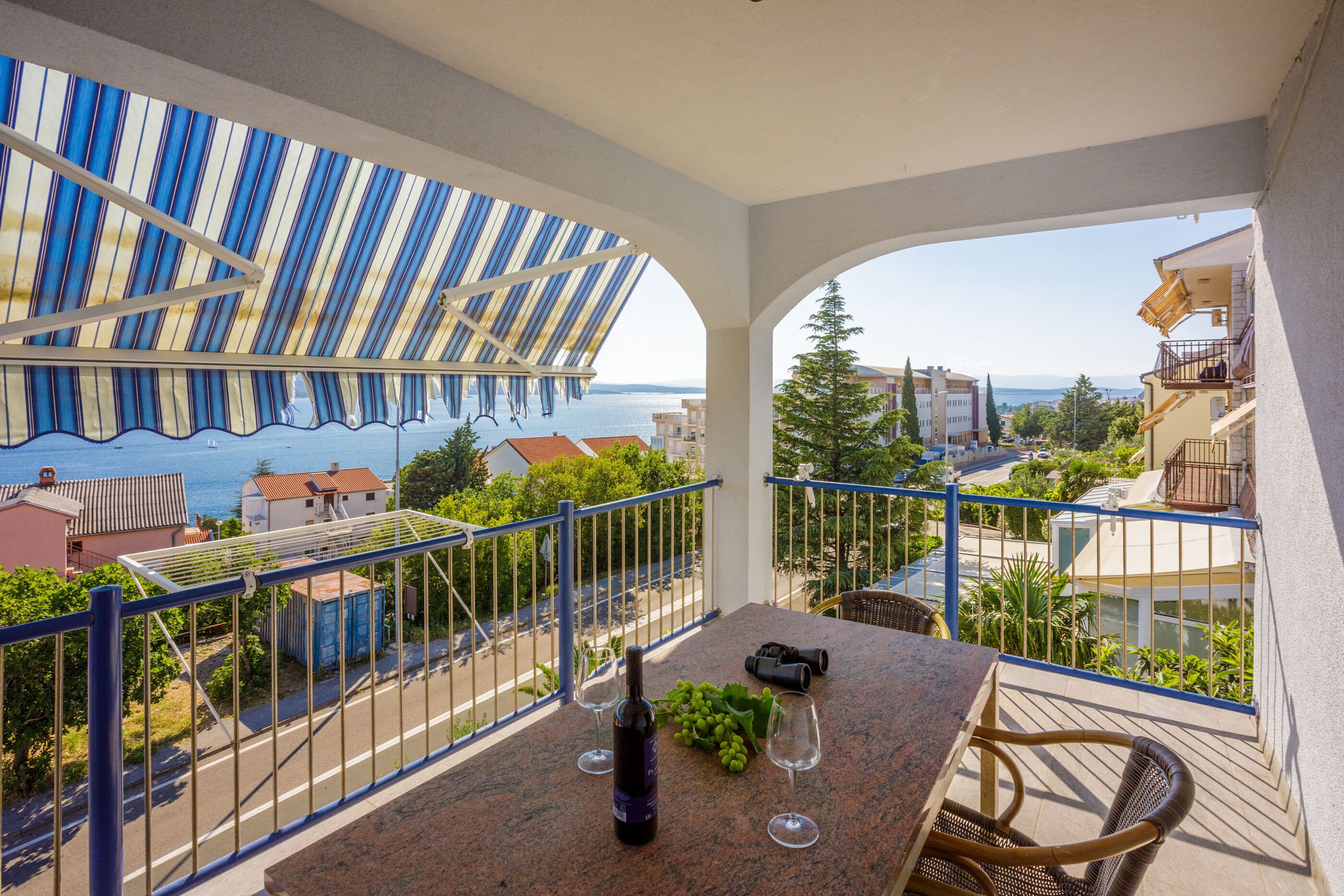 Un balcón con vistas al mar, una mesa con una botella y uvas, y una silla.