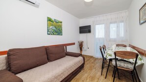 Un salon comprenant un canapé, une table à manger avec des chaises, un téléviseur fixé au mur et une fenêtre avec des rideaux.