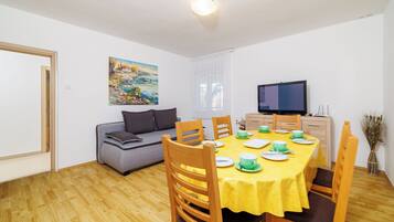 A dining area with a wooden table set for four, a grey sofa, a painting on the wall, and a television on the wall.