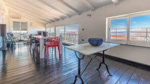 A spacious room with a high ceiling, wooden flooring, a dining table with chairs, and large windows offering a view of the landscape. - Apartments Adriatic (Crikvenica)