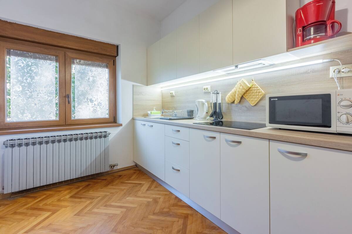 Une cuisine moderne avec des meubles blancs, un parquet en bois disposé en chevrons, un four à micro-ondes, une bouilloire et une machine à café rouge.