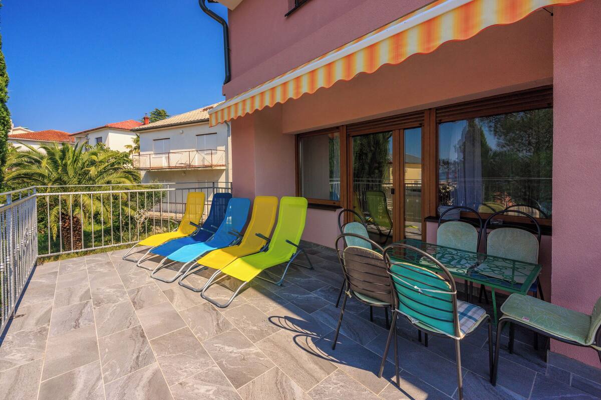 Une terrasse avec des fauteuils colorés et une table, une rambarde et un bâtiment à la façade rose.