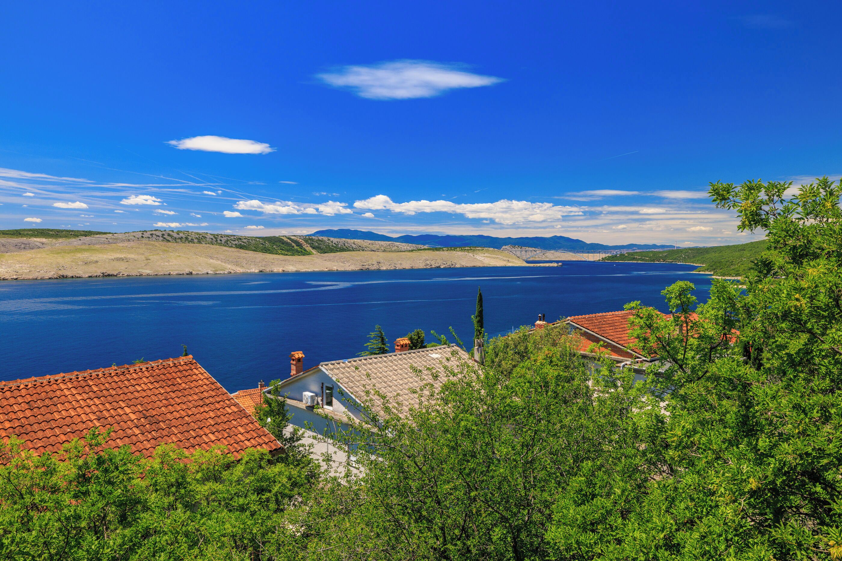 A coastal landscape with a clear view of the sea, a hilly terrain, and a building with a red-tiled roof.