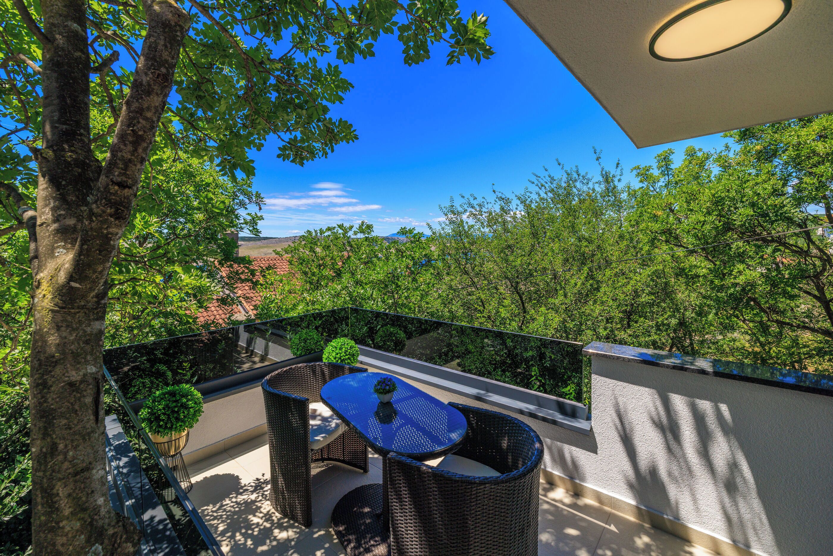 A balcony with wicker furniture, a potted plant, and a view of trees and buildings.