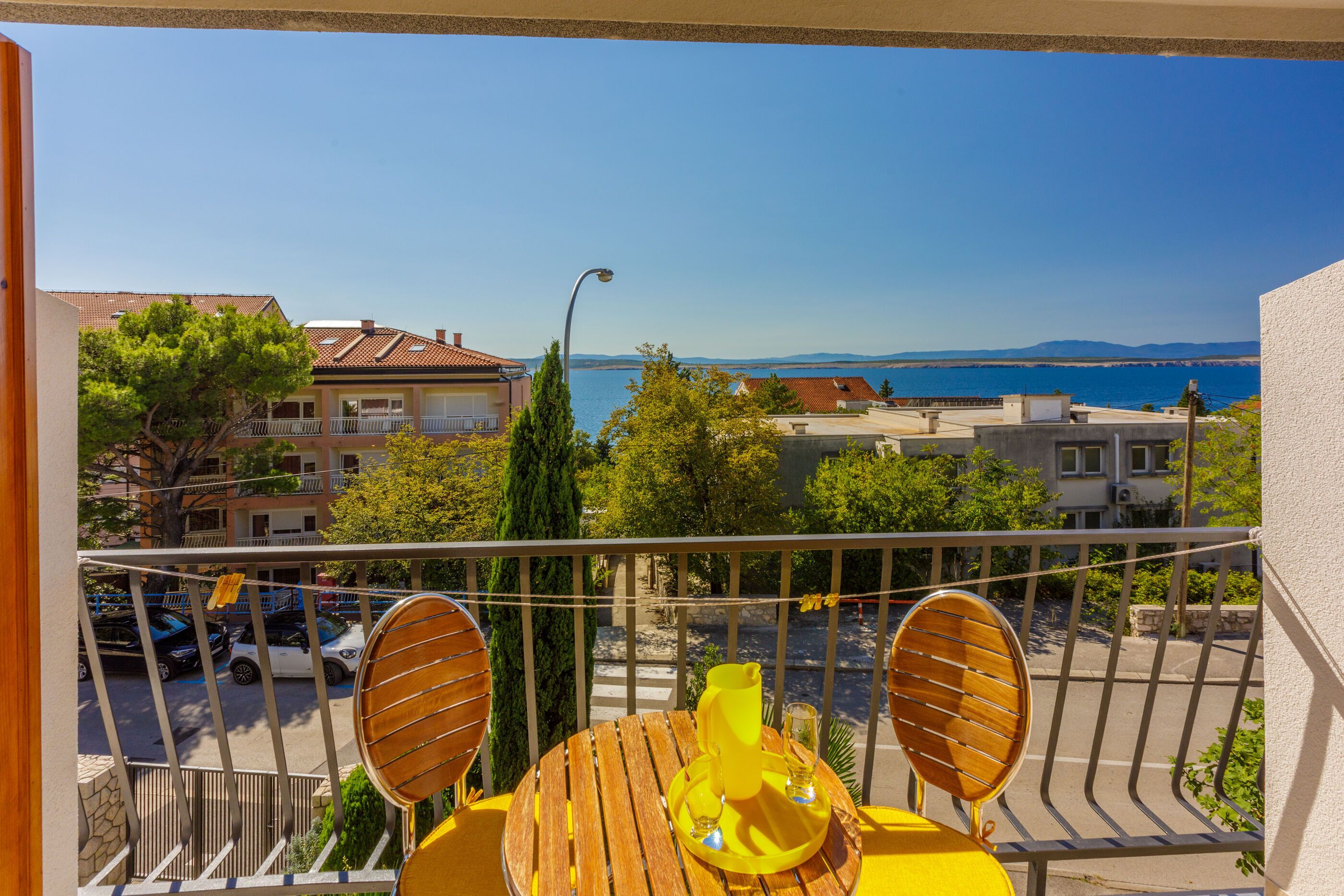 A balcony with a table and chairs, overlooking a coastal area with buildings and a clear sky.