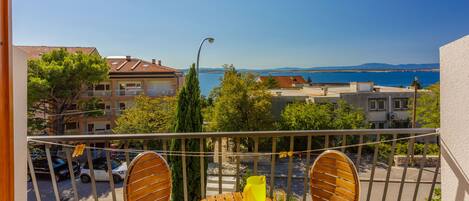 A balcony with a table and chairs, overlooking a coastal area with buildings and a clear sky.
