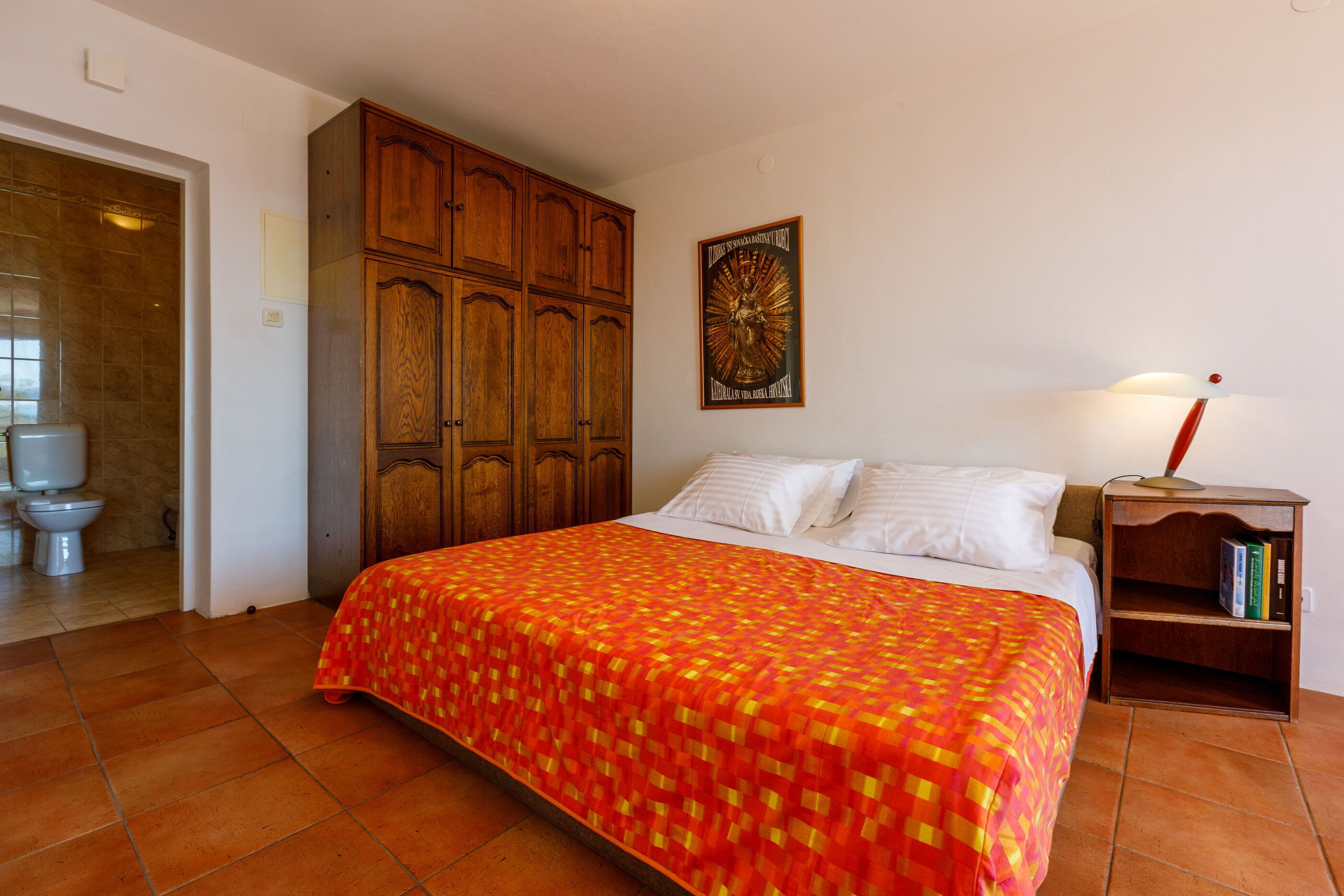 A bedroom with a large bed, wooden wardrobe, and a nightstand with a lamp and books.