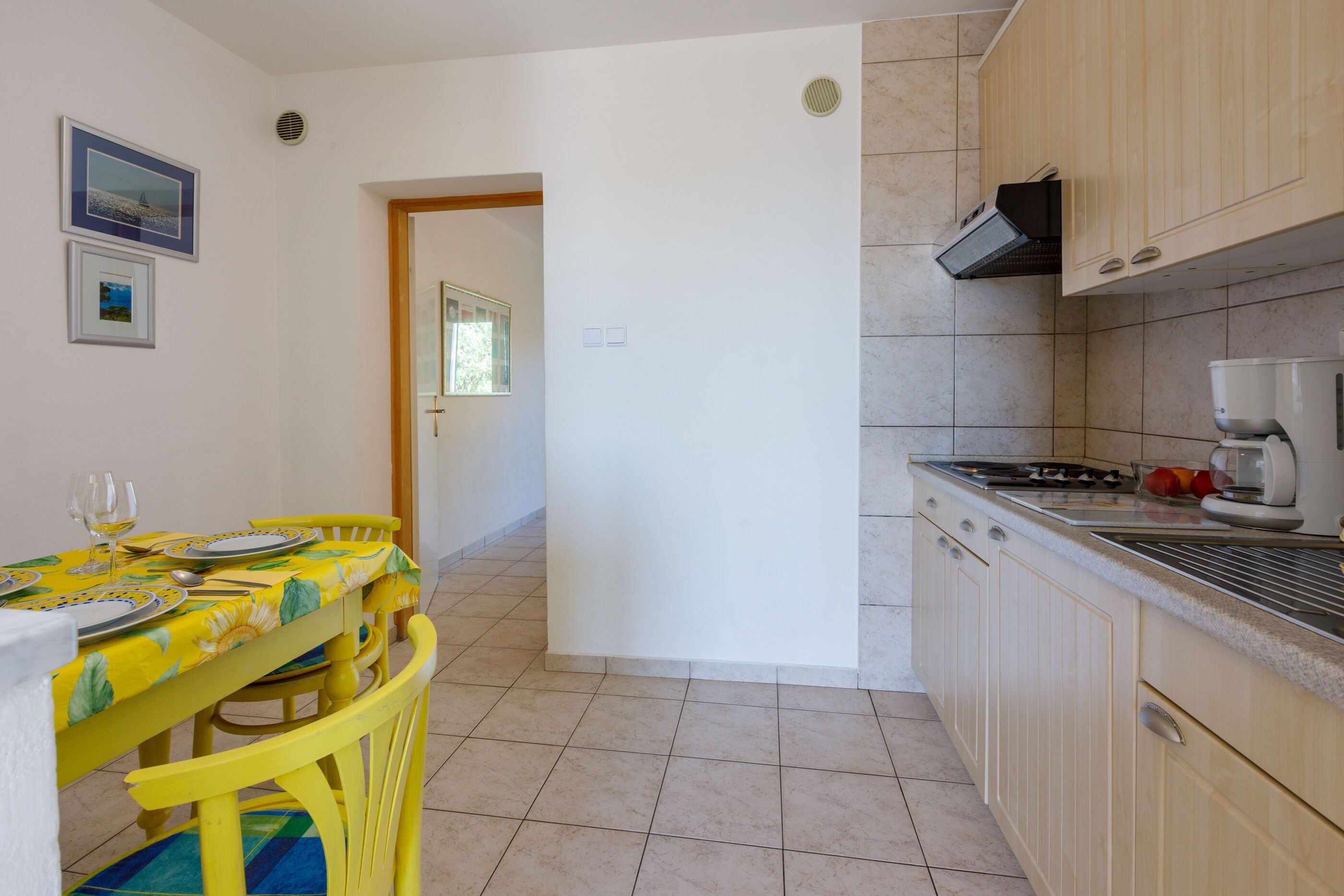 A kitchen with a dining area, featuring a tiled floor, wooden cabinets, and a table set for two with a yellow tablecloth.