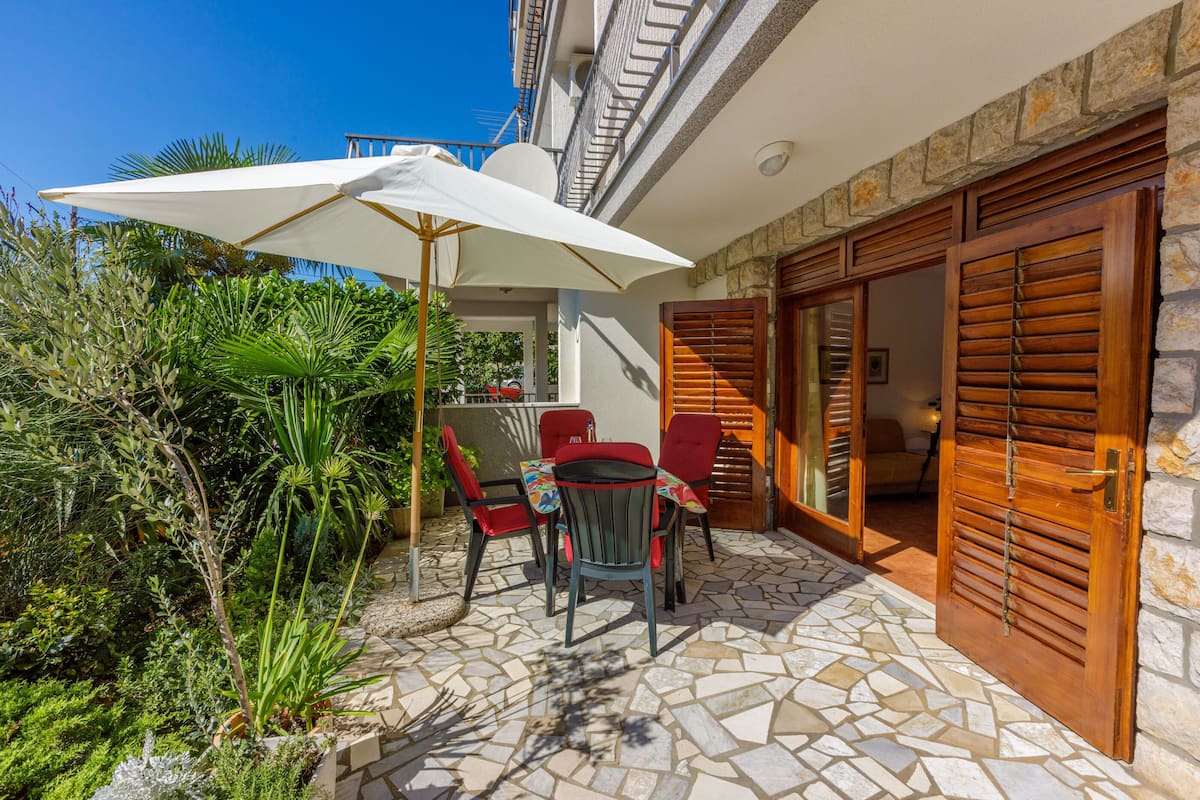 Un patio al aire libre con suelo de piedra, una mesa con sillas, un paraguas blanco y contraventanas de madera.