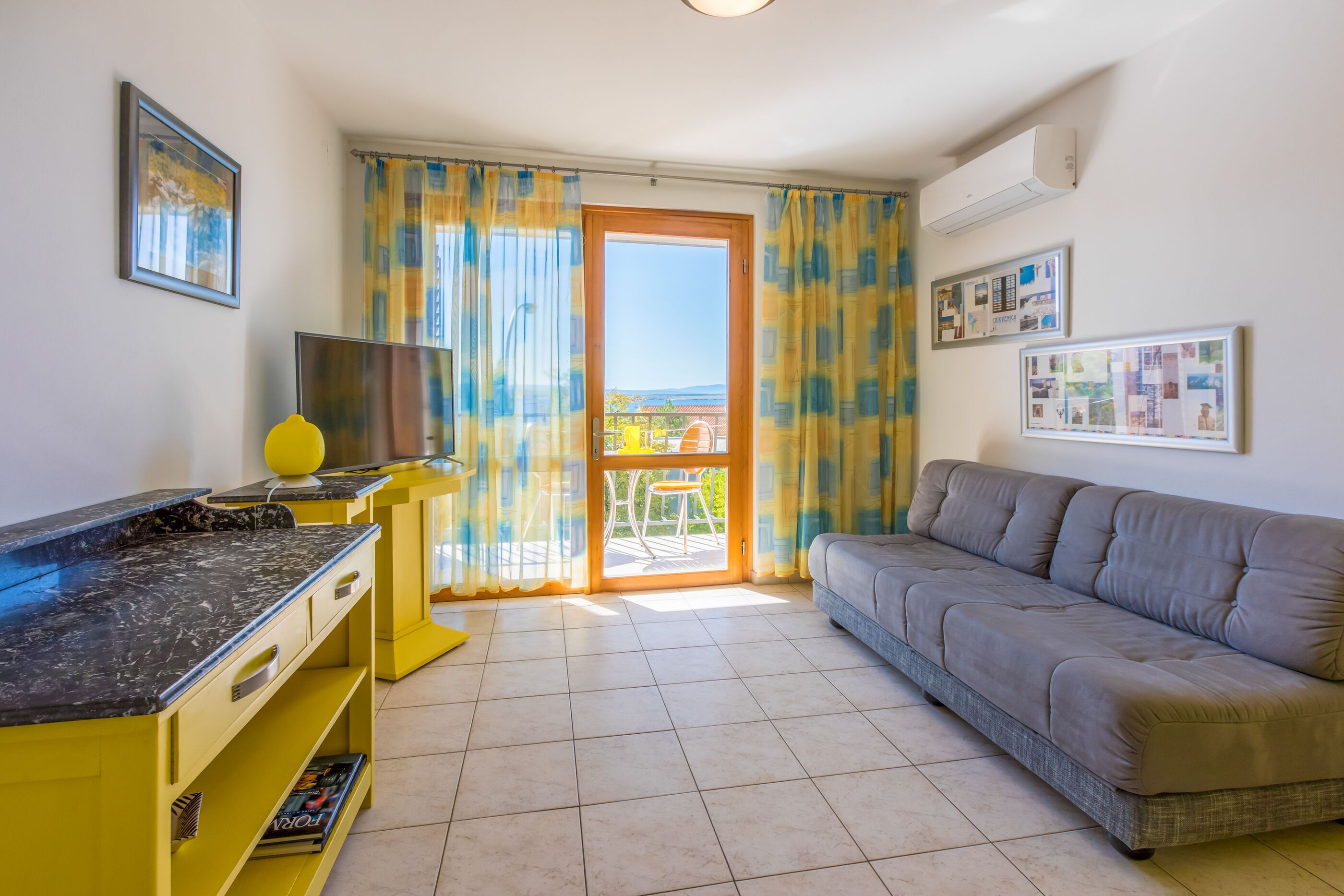 Un salon avec un canapé, un téléviseur, un meuble jaune et un balcon avec des rideaux.