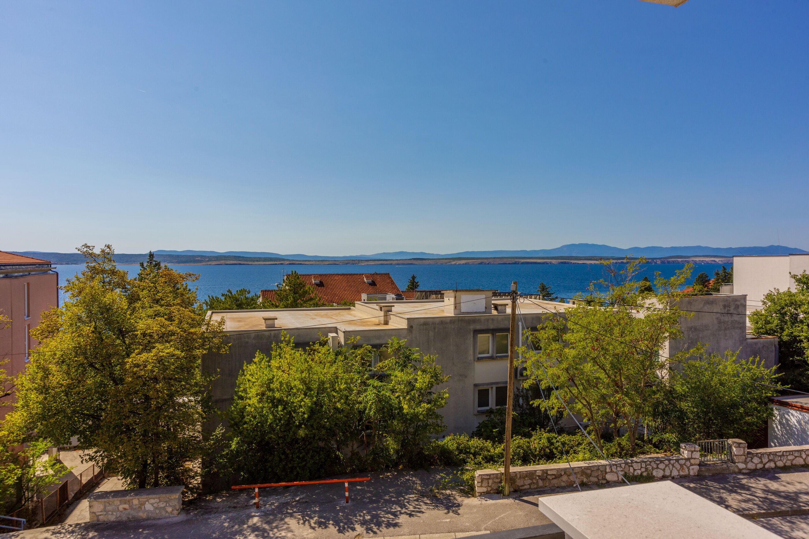 A coastal residential area with buildings, trees, and a clear view of the sea and distant mountains.