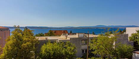 A coastal residential area with buildings, trees, and a clear view of the sea and distant mountains.