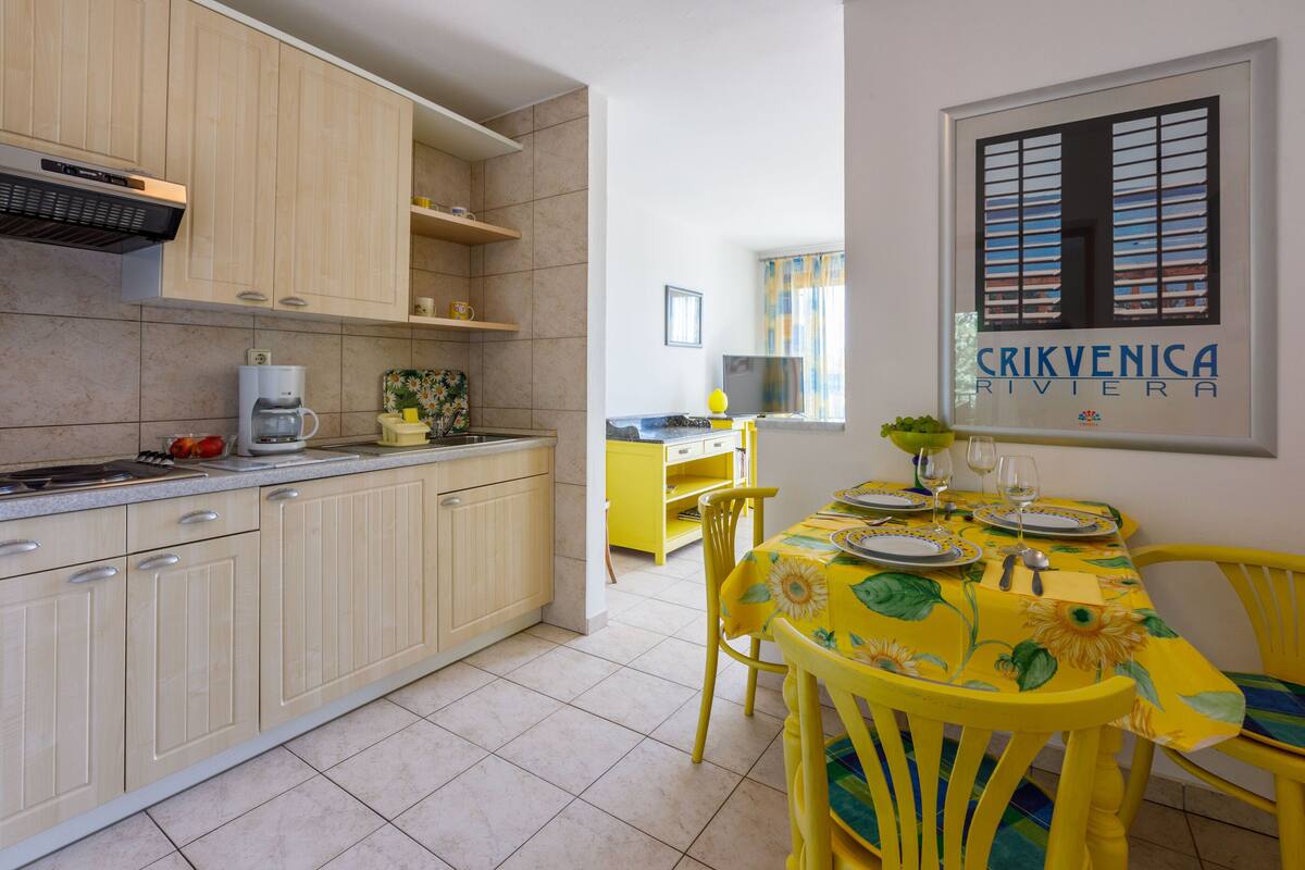 Una cocina con armarios de madera clara, una cafetera y un comedor con sillas amarillas y una mesa dispuesta para dos.
