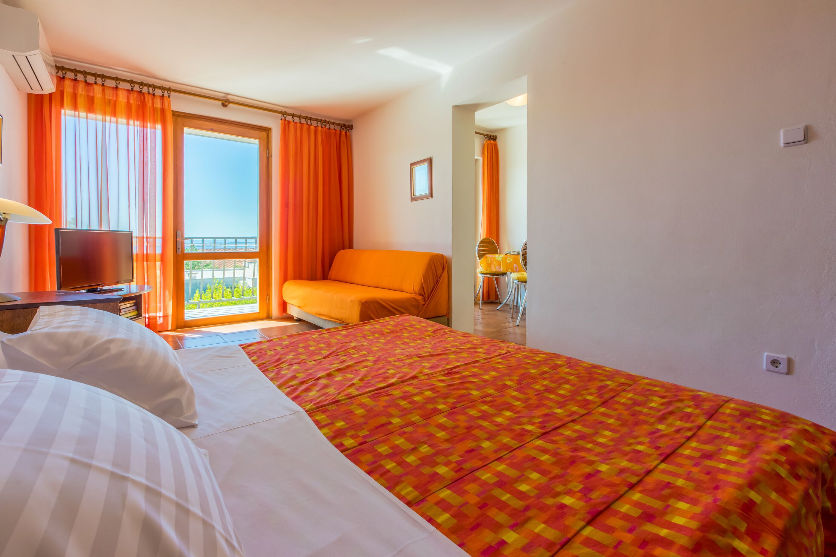 Ein Hotelzimmer mit einem großen Bett, einer Couch, einem Fernseher und einem Balkon mit orangenen Vorhängen.
