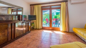 Una habitación con un armario de madera, televisor, sofá amarillo y puerta corrediza de cristal que da a un espacio exterior con sillas y una mesa.