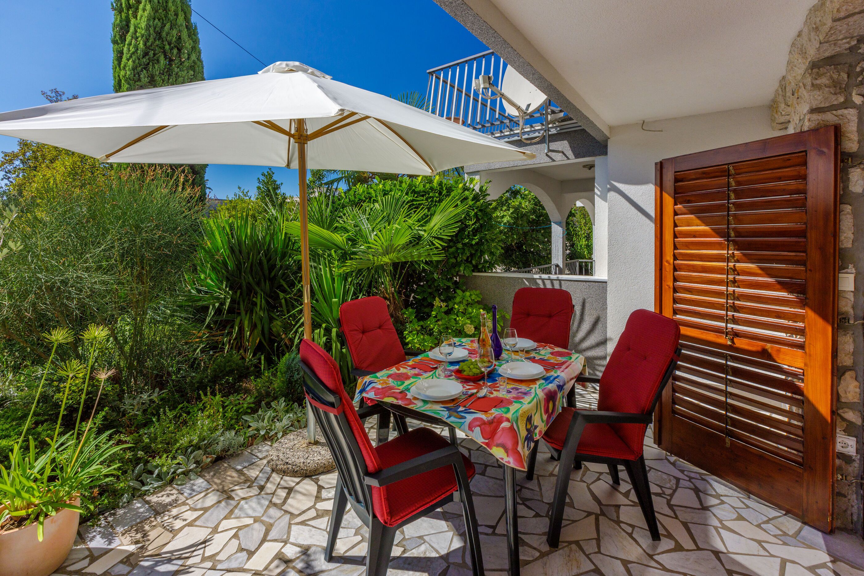 Une terrasse extérieure avec une table dressée pour quatre personnes, entourée de chaises rouges, d’un parasol blanc et d’un sol carrelé en pierre.
