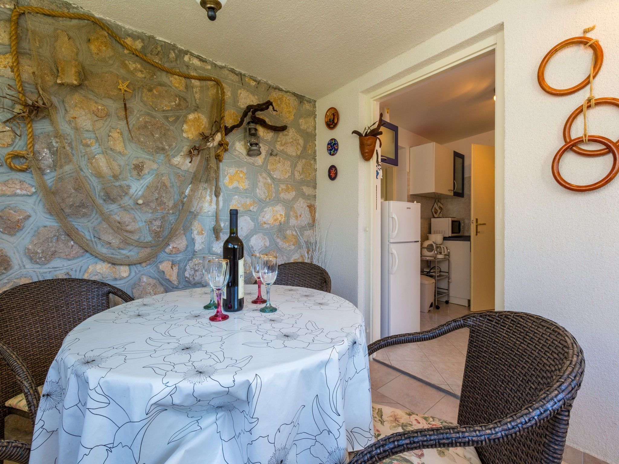 A dining area with a round table covered by a floral tablecloth, surrounded by wicker chairs. A bottle of wine and two glasses are on the table. The wall behind features a stone pattern and a decorative rope.