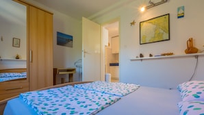 Ein Schlafzimmer mit einem Bett, einem Holzschrank, einem kleinen Tisch und einem durchgehenden Türrahmen erkennbaren Küchenbereich.