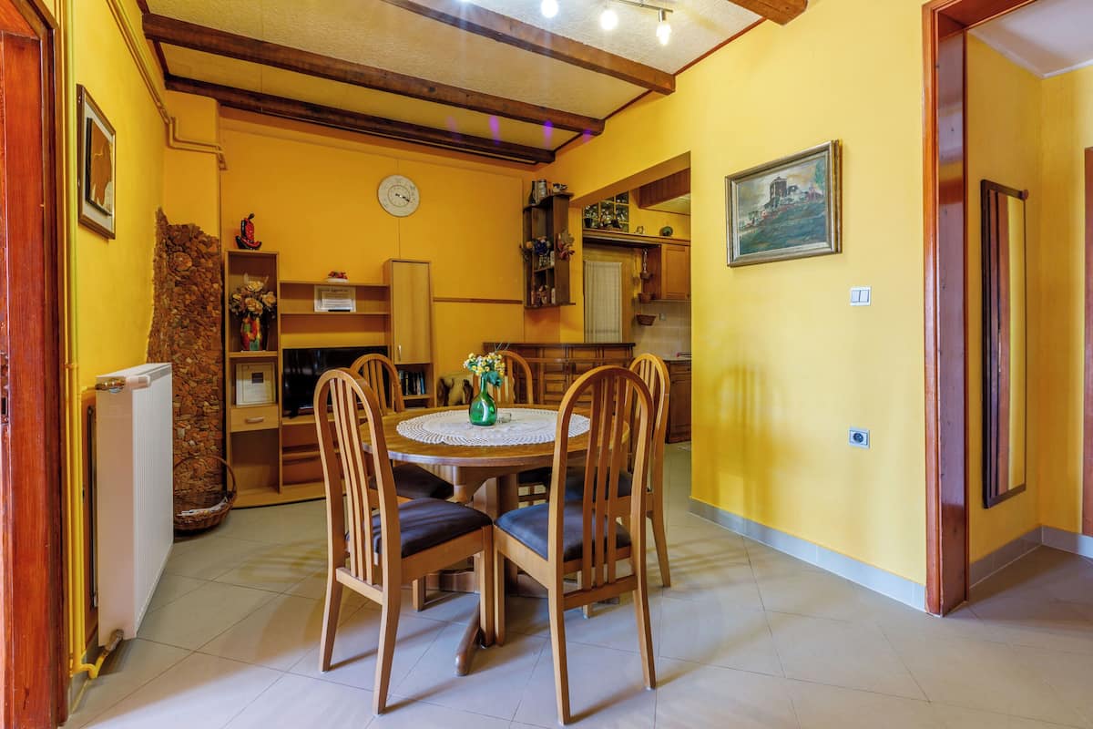 A dining area with a round table and chairs, a clock on the wall, and a kitchen in the background.