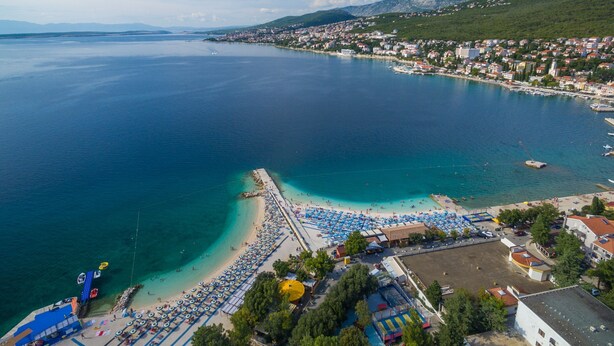 An aerial view of a coastal town with a beach, clear blue waters, and a mix of residential and commercial buildings. - Apartment Giardino (Selce)