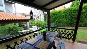 A covered patio with a wooden table and benches, a potted plant, and a view of a residential area with a green hedge and a parked car. - Apartment Giardino (Selce)