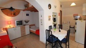 A compact hotel room with a bed, a small desk, a TV, a dining table with chairs, and a kitchen area with a fridge and a coffee maker. - Apartment Giardino (Selce)