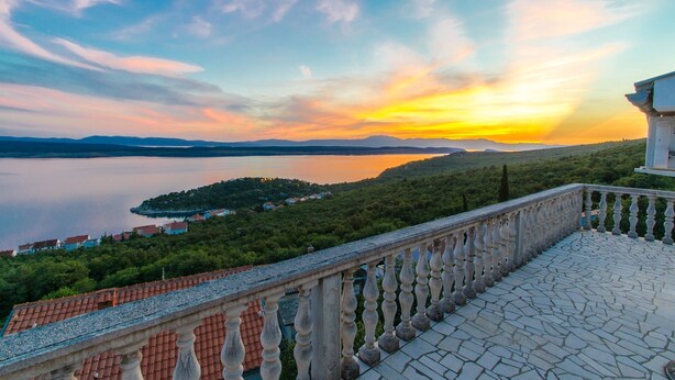 Un balcon doté d’une balustrade en pierre, offrant une vue sur un lac et un coucher de soleil.