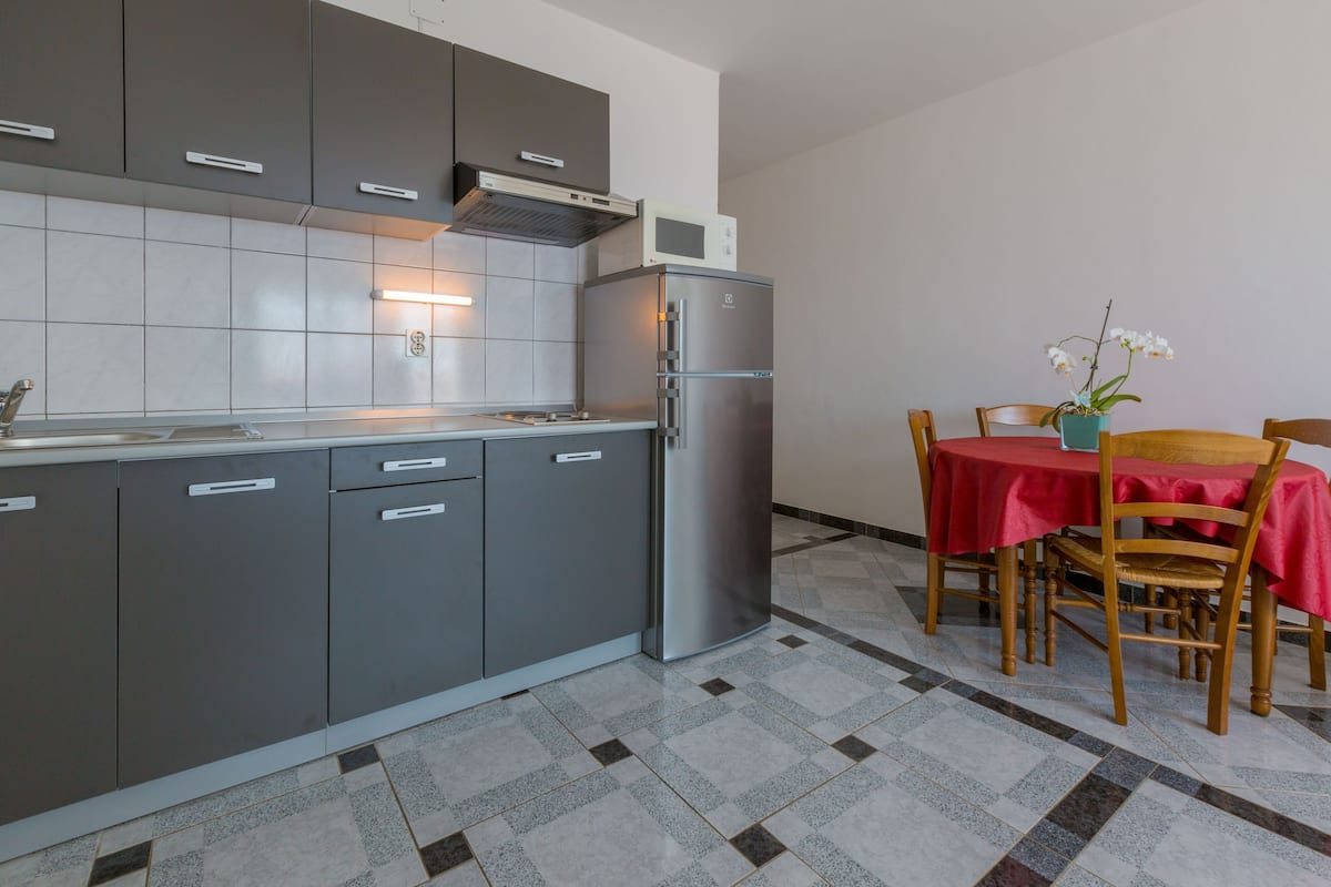 A modern kitchen with grey cabinets, a stainless steel refrigerator, and a dining area with a red tablecloth and chairs.