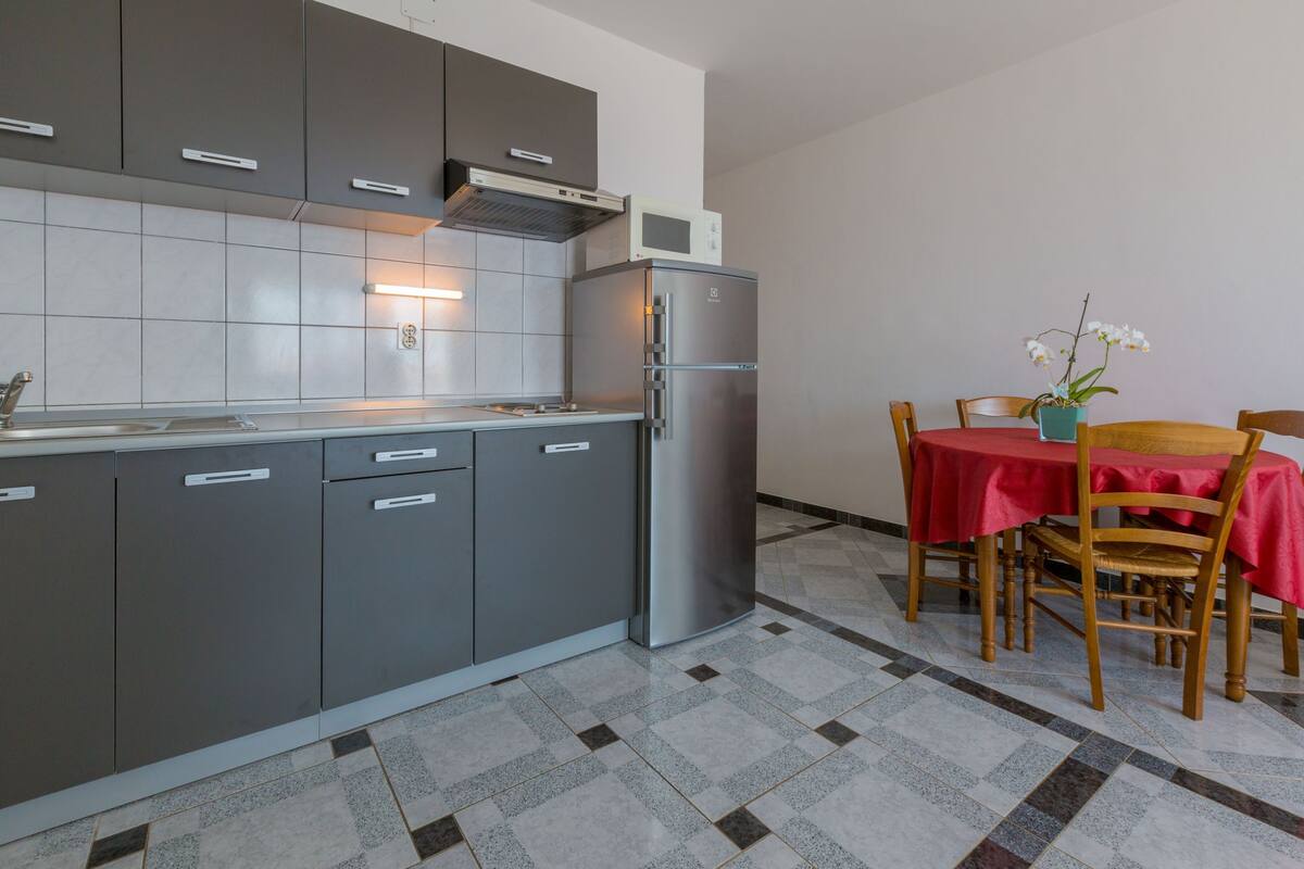 Una cocina moderna con armarios grises, un refrigerador de acero inoxidable y una zona de comedor con mantel y sillas rojos.