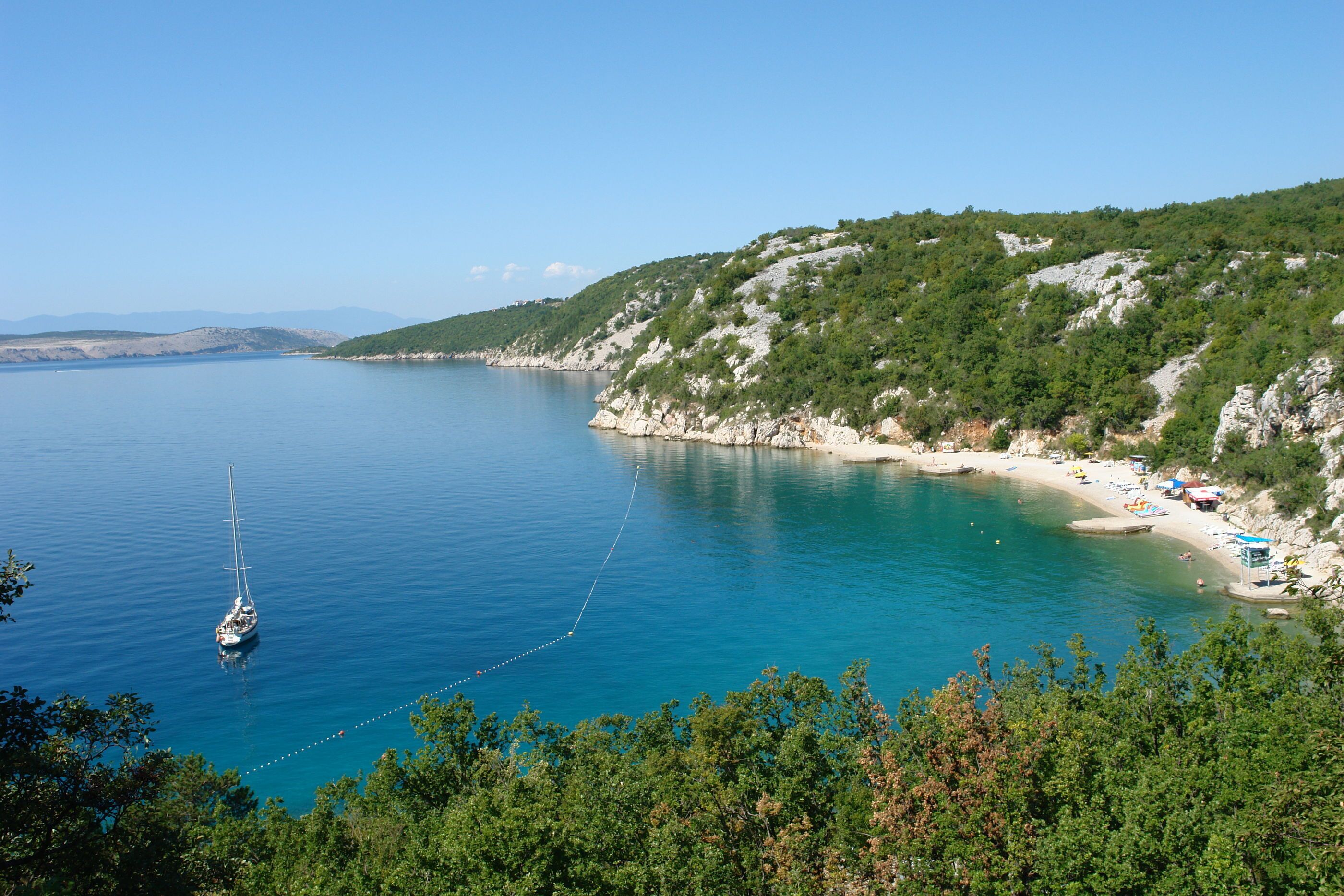 Eine Küstenlandschaft mit blauem Meer, einem Segelboot, das in der Nähe des Ufers vor Anker liegt, und einem Strand mit Personen und Sonnenschirmen.