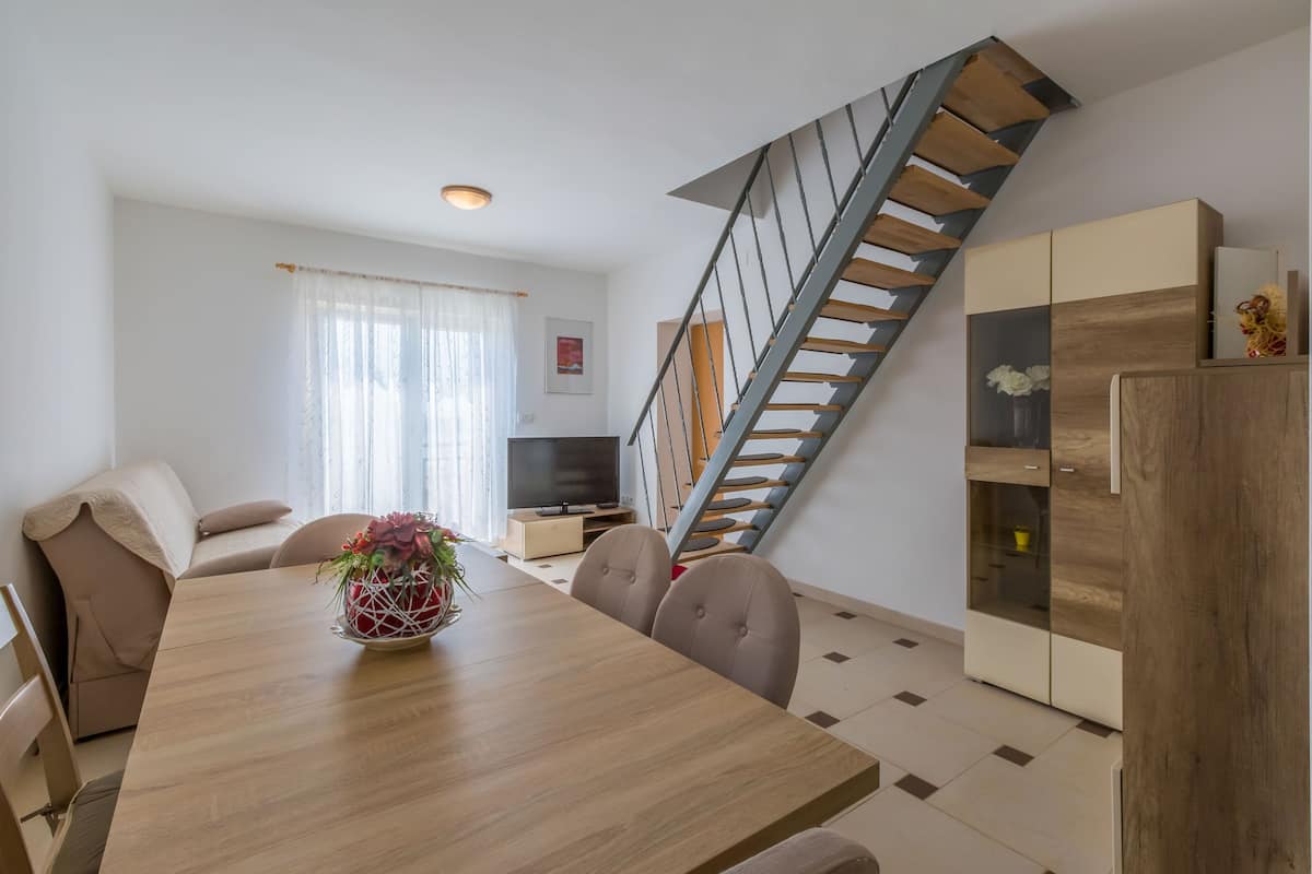 A modern living room with a wooden dining table, beige sofa, and a staircase with a wooden railing.